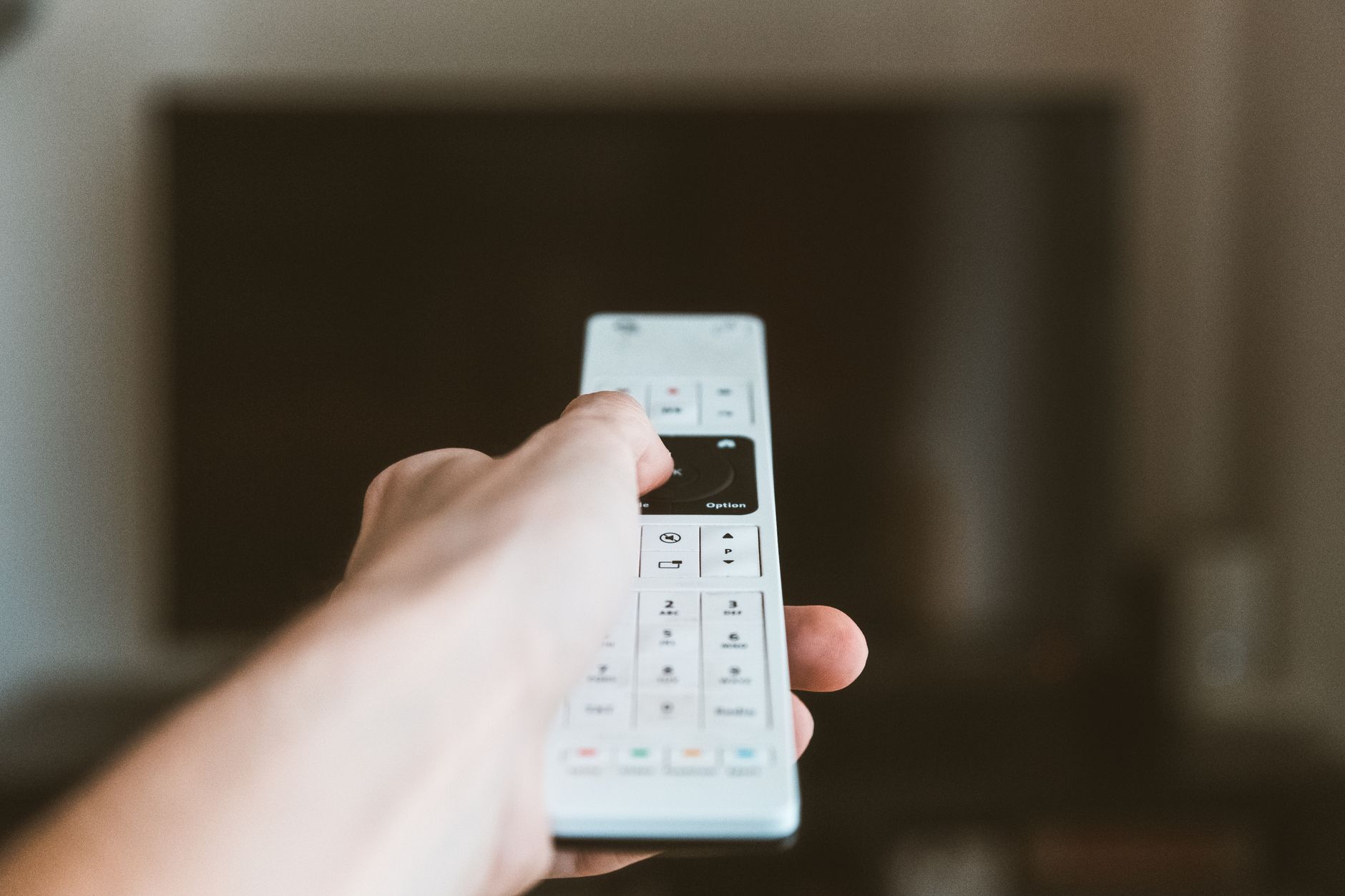 A hand holding an Apple TV Siri Remote pointed toward a living room television screen.